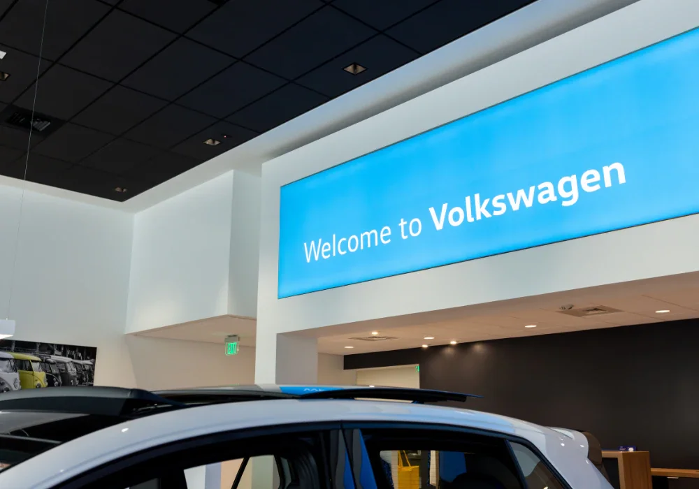 A brightly lit 'Welcome to Volkswagen' sign on a blue banner inside a modern dealership, with a white car in the foreground.