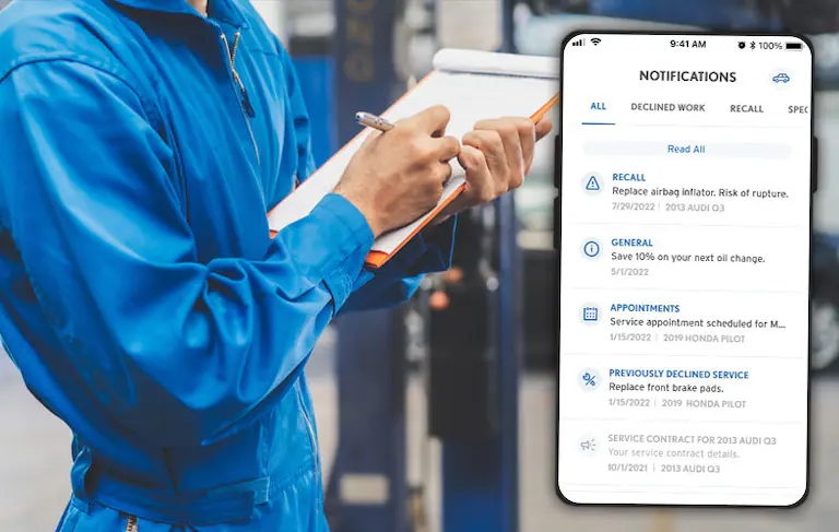 Mechanic writing on clipboard next to phone displaying car service notifications app.
