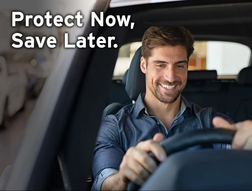 A smiling man in a car, hands on the steering wheel, with 'Protect Now, Save Later.' text overlaid.