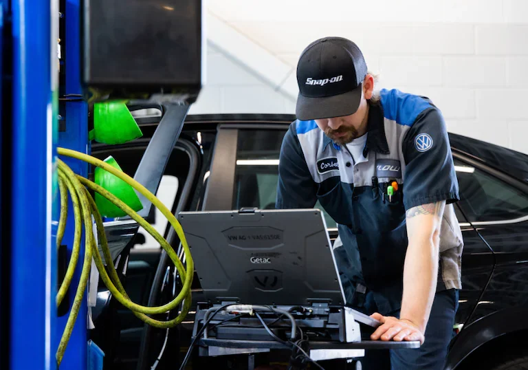 A male mechanic in uniform and a Snap-on hat works on a laptop in a service bay, a black car behind him.