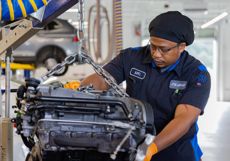 A mechanic named Karl wearing glasses and a dark blue uniform carefully lowers a car engine with a hoist.