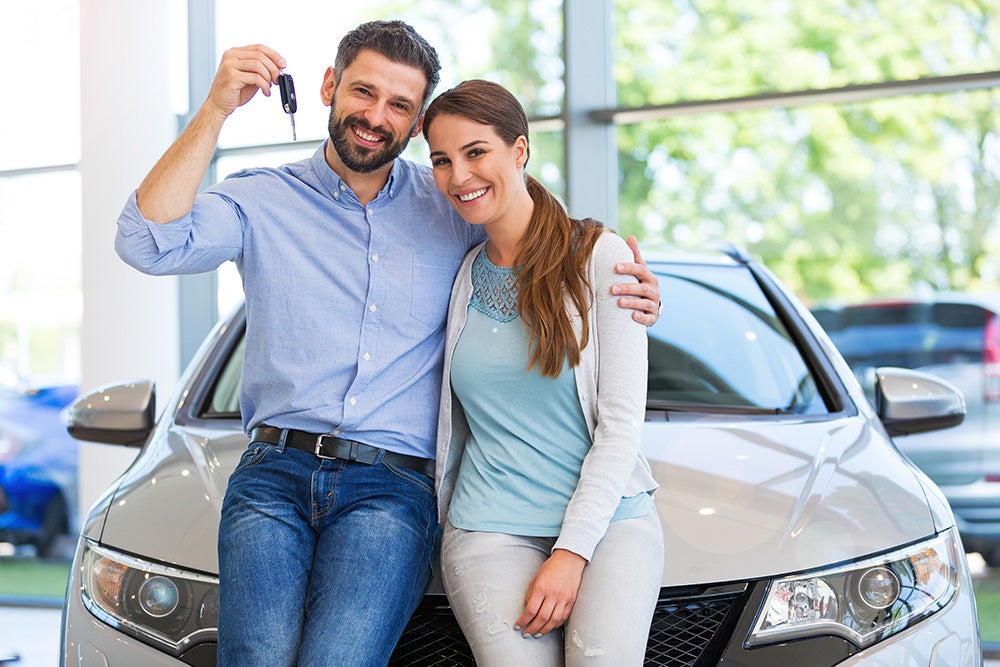 A smiling couple embracing beside a new silver car in a showroom, man holding up car keys.