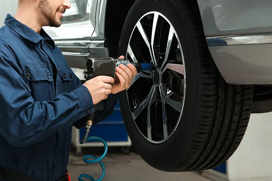 Mechanic in blue overalls using an impact wrench to change a tire on an elevated gray car.
