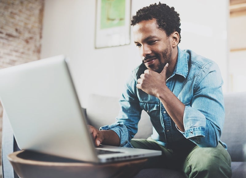 A smiling man with a beard sits on a sofa, looking at a laptop screen with his hand on his chin.